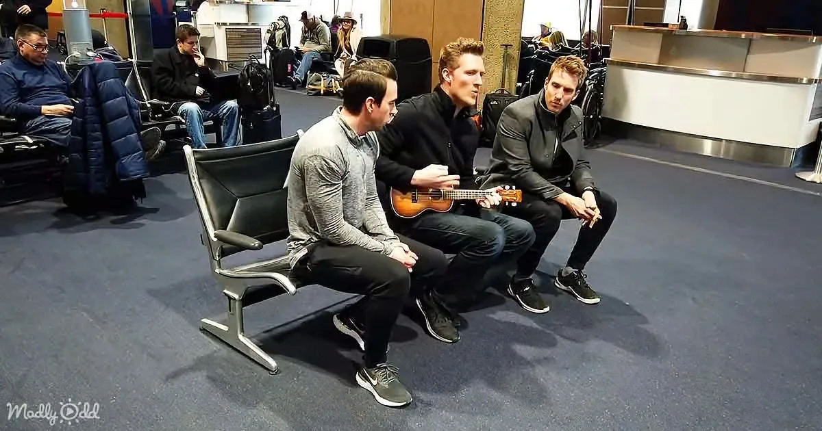 Three men sit on a bench in an airport, one playing a ukulele while others watch.
