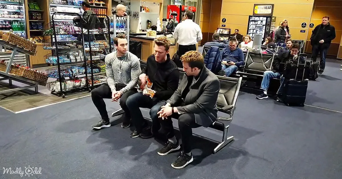 Three men sit on a bench in an airport lounge, one playing guitar, while others wait with luggage.