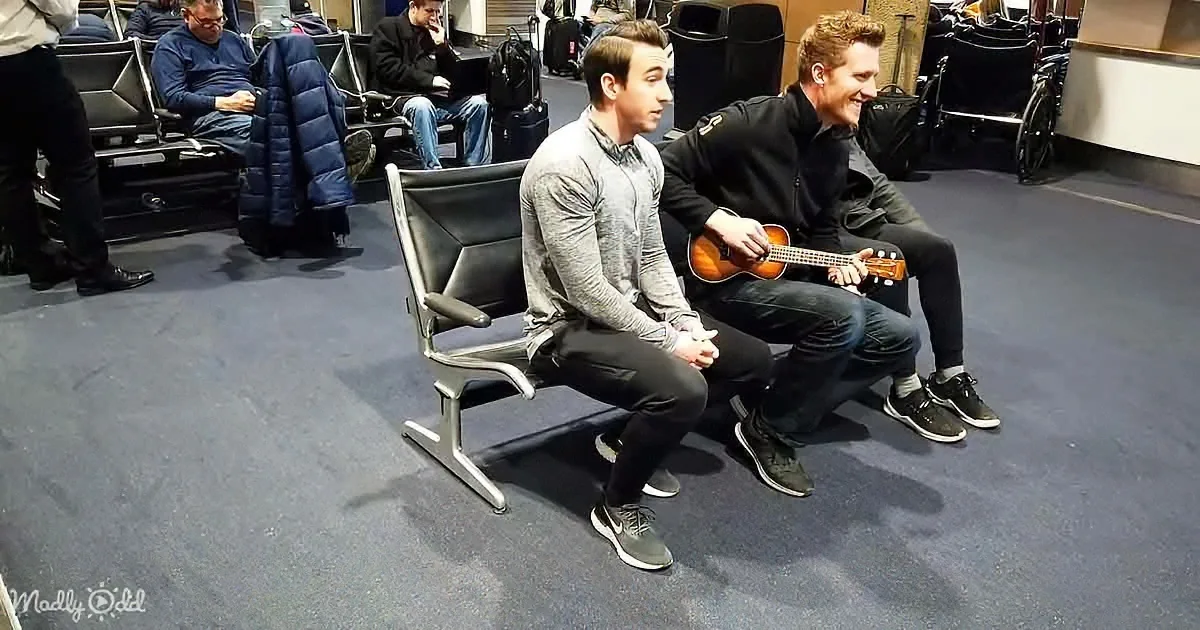 Two men sit on a bench in an airport, one playing a ukulele while the other watches.