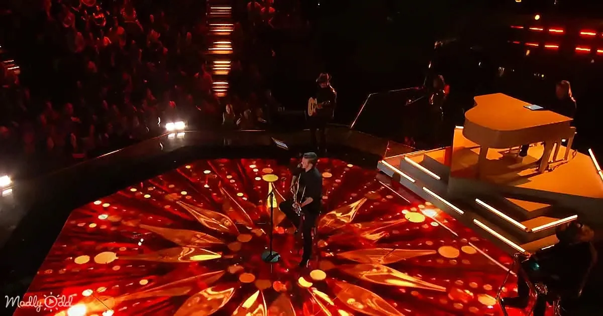 A performer stands on a vibrant, illuminated stage with a red and orange pattern, surrounded by a dimly lit audience and...