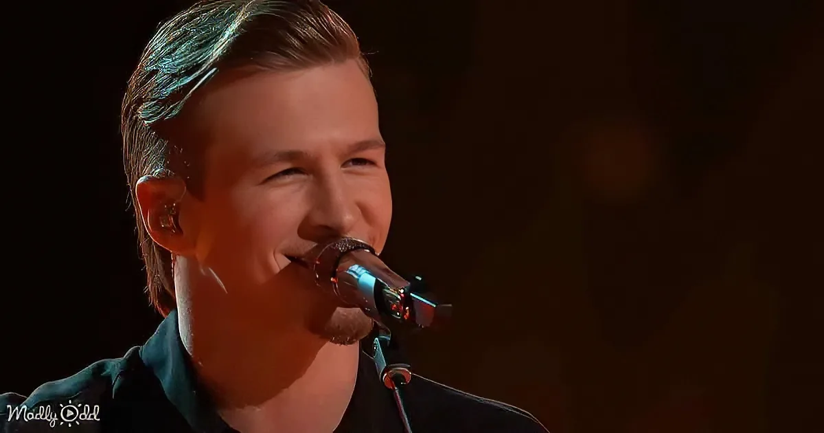 A person singing into a microphone, wearing a dark shirt, with a blurred background.