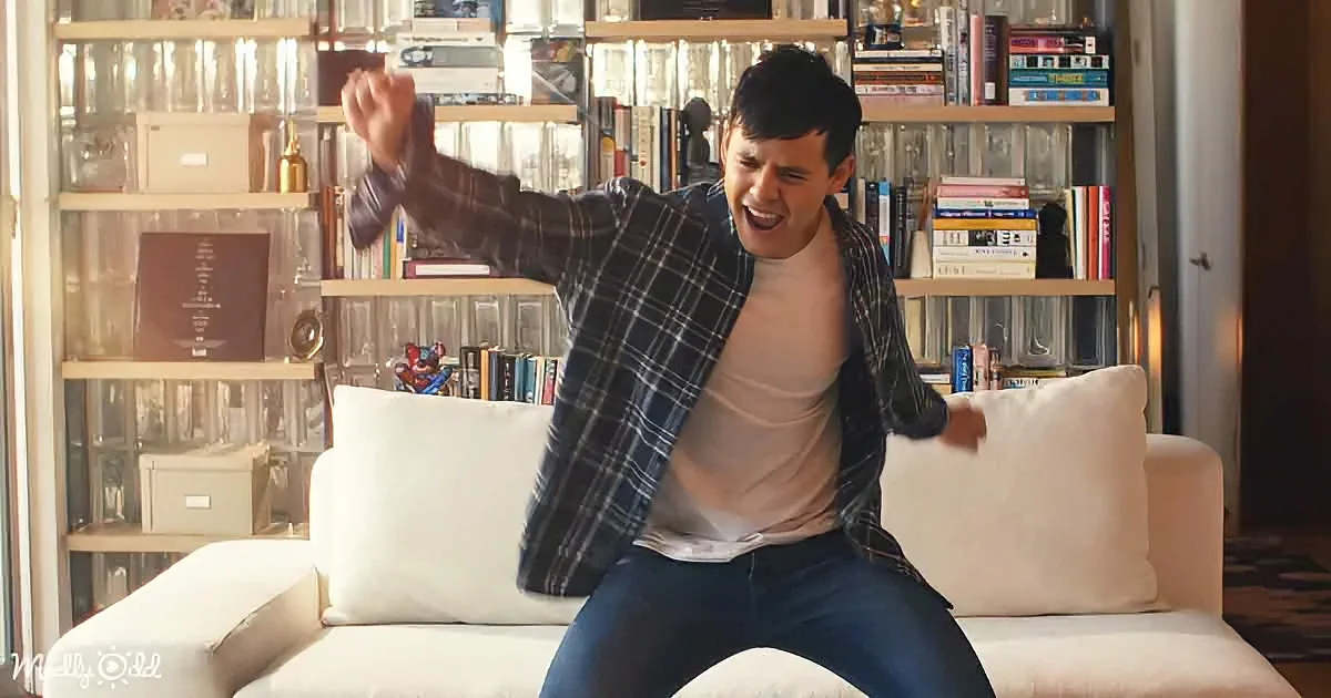 A man in a plaid shirt and jeans is sitting on a couch, raising his arms in excitement.