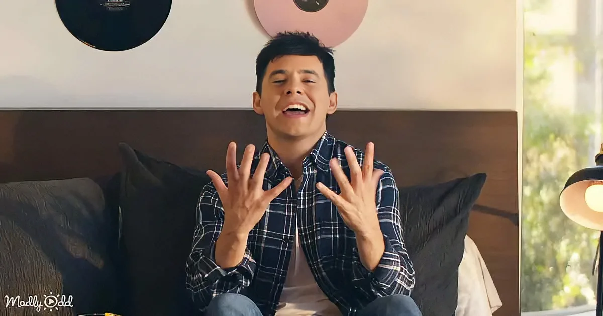 A man in a plaid shirt sits on a couch, smiling widely with his hands raised, displaying all ten fingers.
