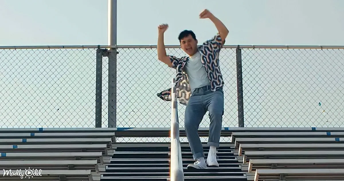 A person stands on bleachers, arms raised, wearing a patterned shirt and jeans, with a chain-link fence and sky in the...