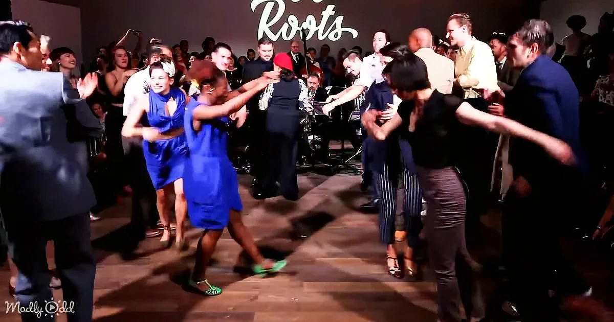 A group of people are dancing energetically on a wooden floor, with a band playing in the background.