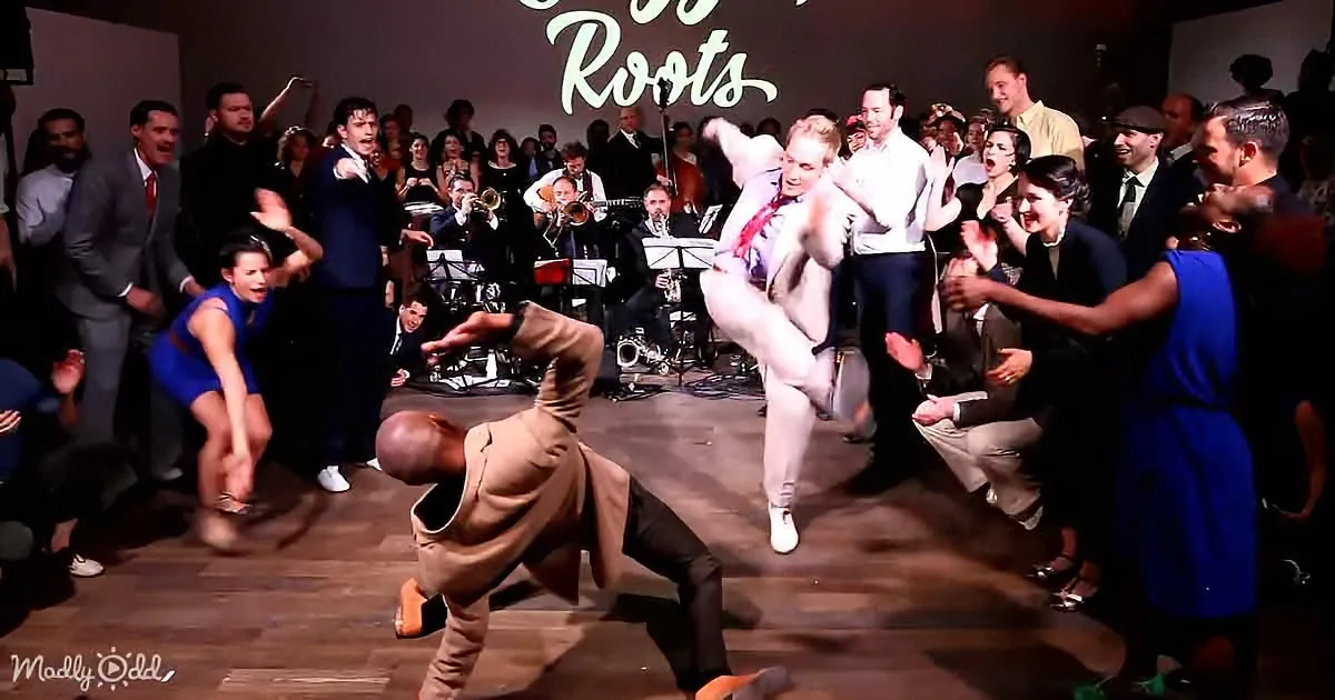 A man in a tan suit is breakdancing on a stage surrounded by an enthusiastic crowd and a band.