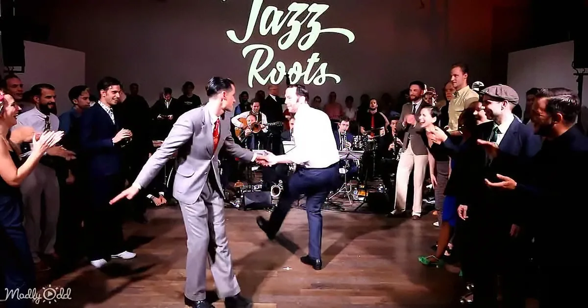 Two men in suits dance in front of a jazz band and audience at a "Jazz Roots" event.