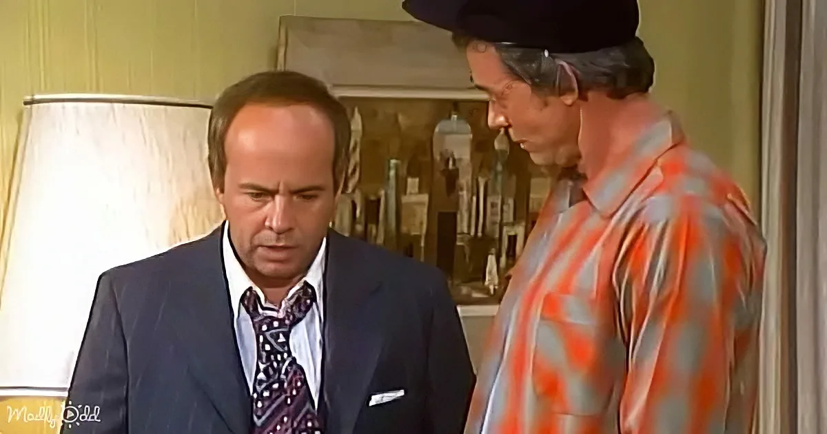 A man in a suit and tie looks down while another man in a striped shirt and hat stands beside him.
