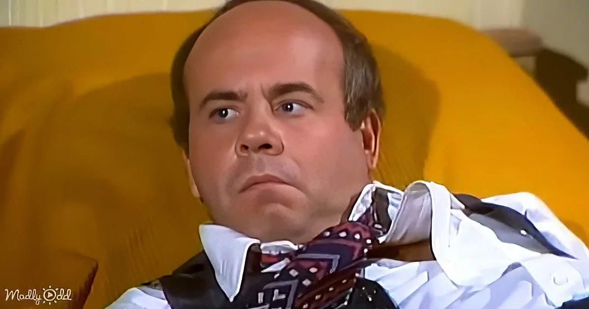 A man in a white shirt and bow tie sits on a yellow couch, looking slightly to the side with a serious expression.
