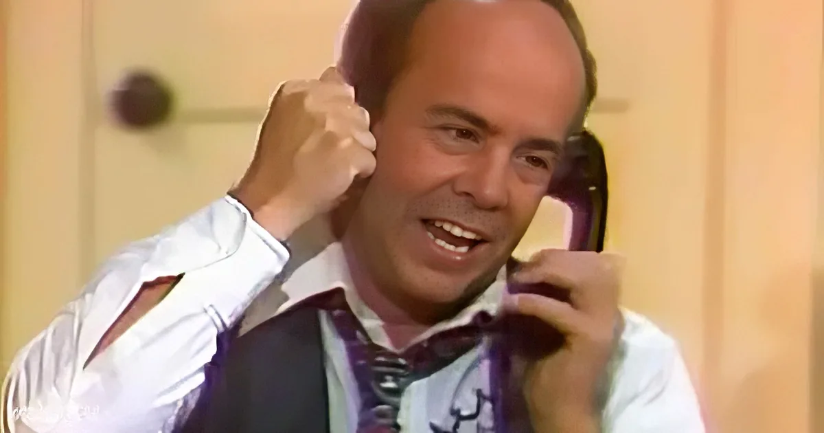 A man in a white shirt and purple tie is holding a phone to his ear with a smile.