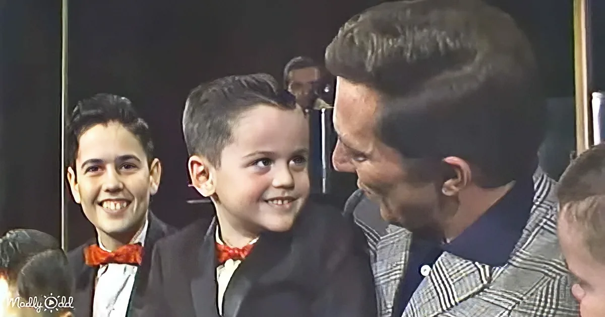 A young boy in a suit smiles at an older man in a checkered suit, surrounded by others in formal attire.