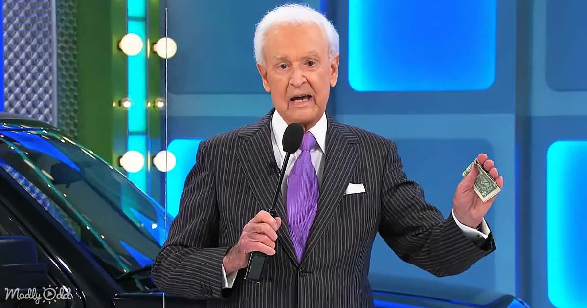 A man in a suit holds a microphone and a dollar bill, standing in front of a car and a colorful backdrop.