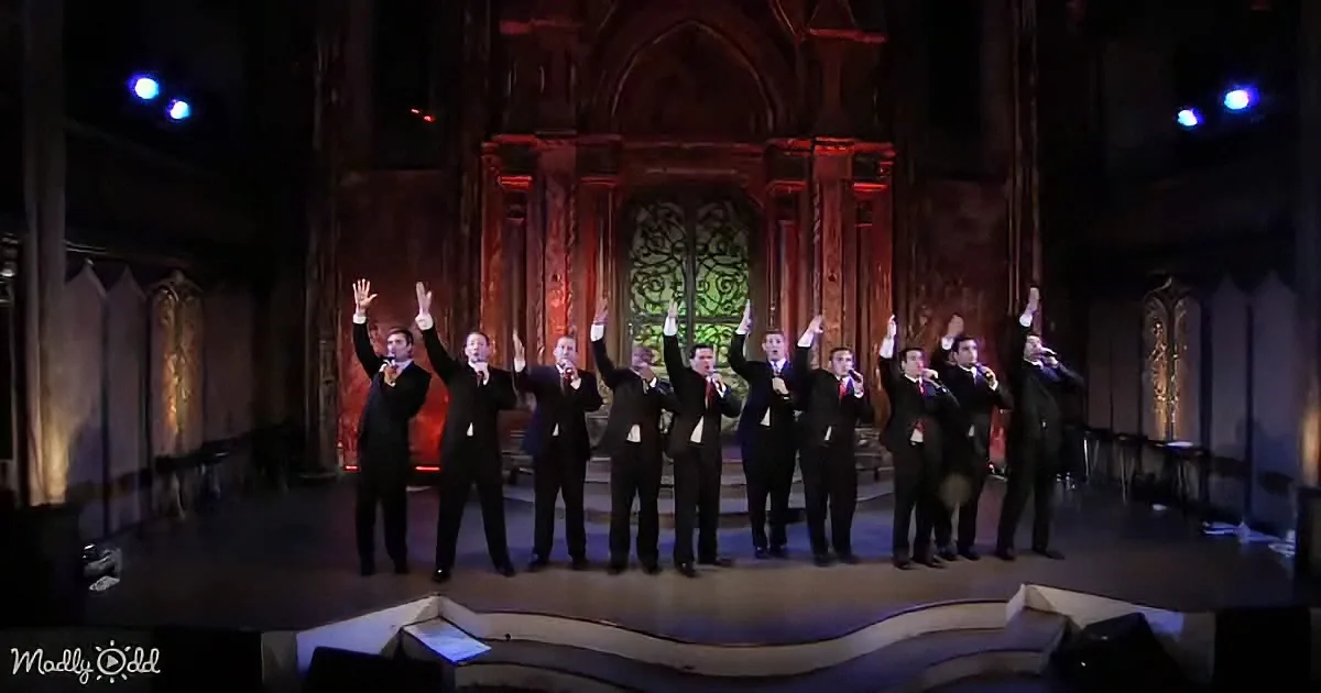 A group of men in black suits sing on stage with their hands raised, illuminated by red and blue lights.