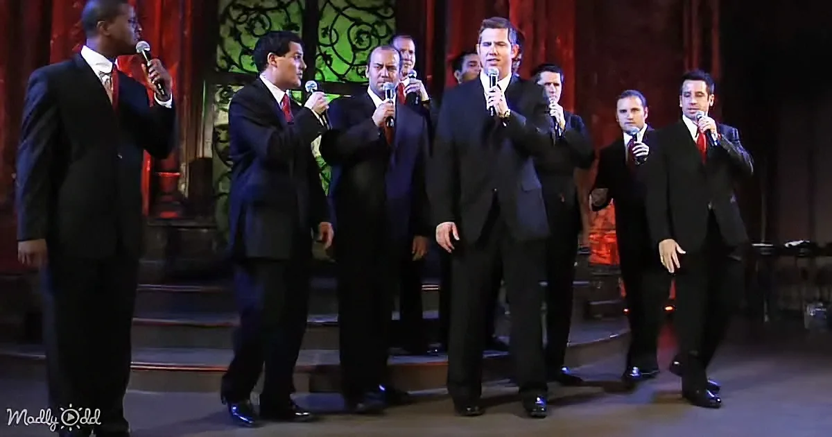 A group of men in suits sing on stage, holding microphones, with ornate red curtains in the background.