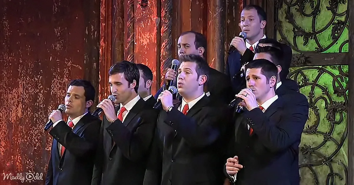 A group of men in suits sing into microphones on stage with ornate background.