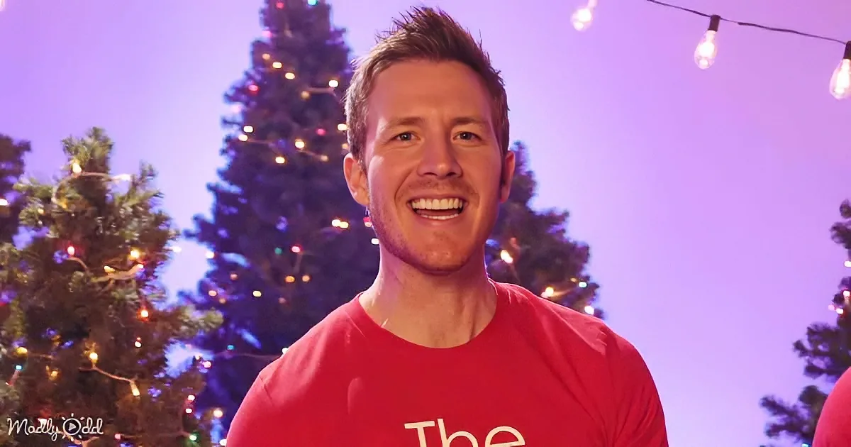 A smiling man in a red shirt stands in front of a Christmas tree with lights, under a purple sky.