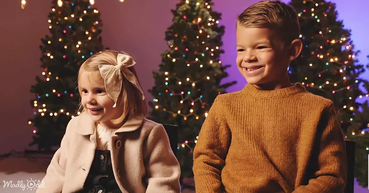 A young boy and girl smile in front of decorated Christmas trees, wearing festive attire.