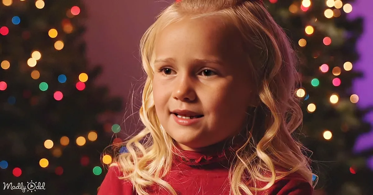 A young girl with blonde hair in a red sweater smiles in front of a Christmas tree with colorful lights.