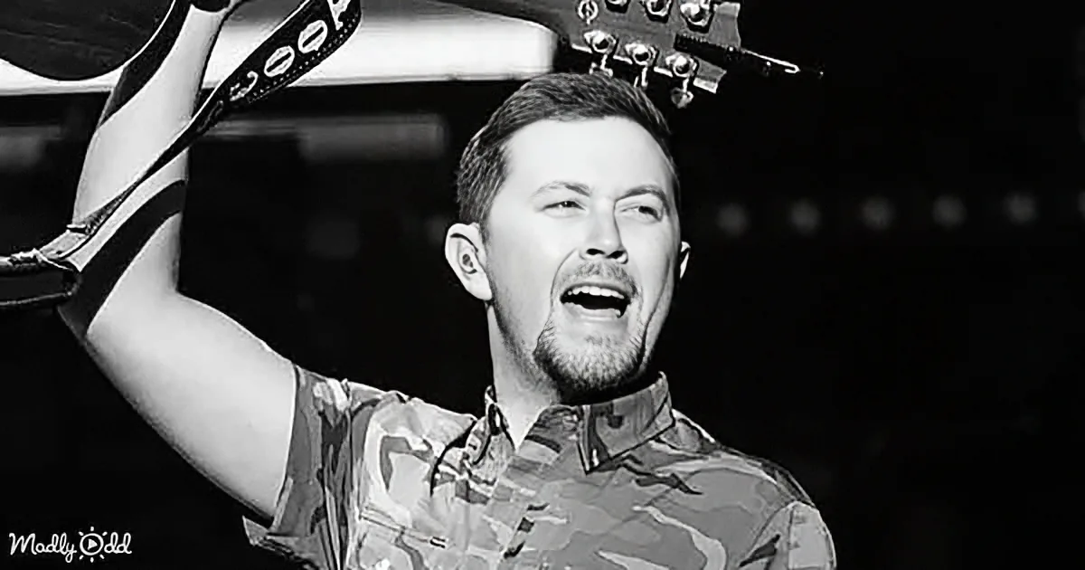 A man in a camouflage shirt holds a guitar strap above his head, singing passionately.