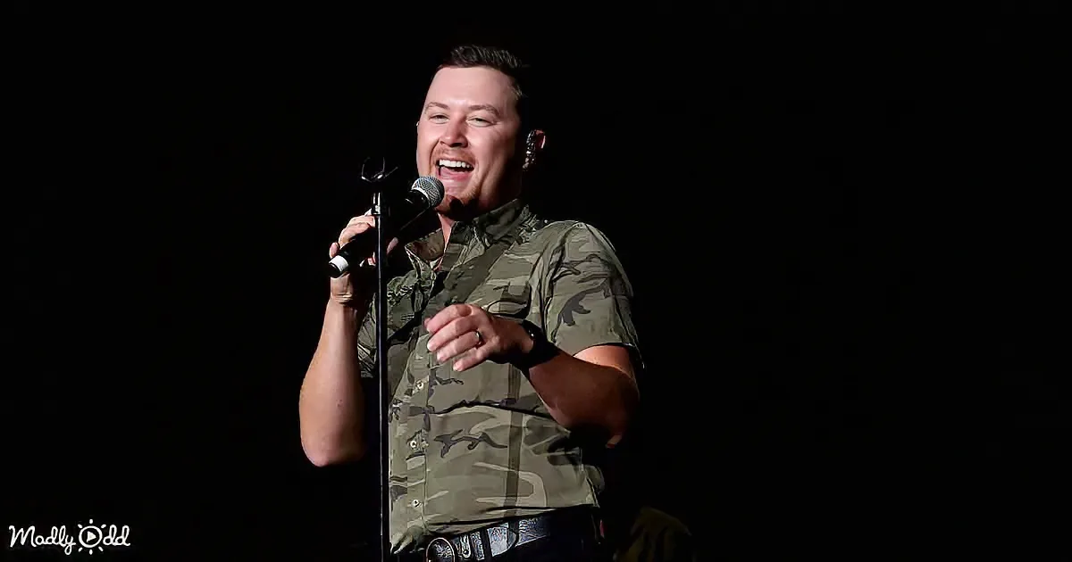A man in a camouflage shirt sings into a microphone on stage, smiling widely.
