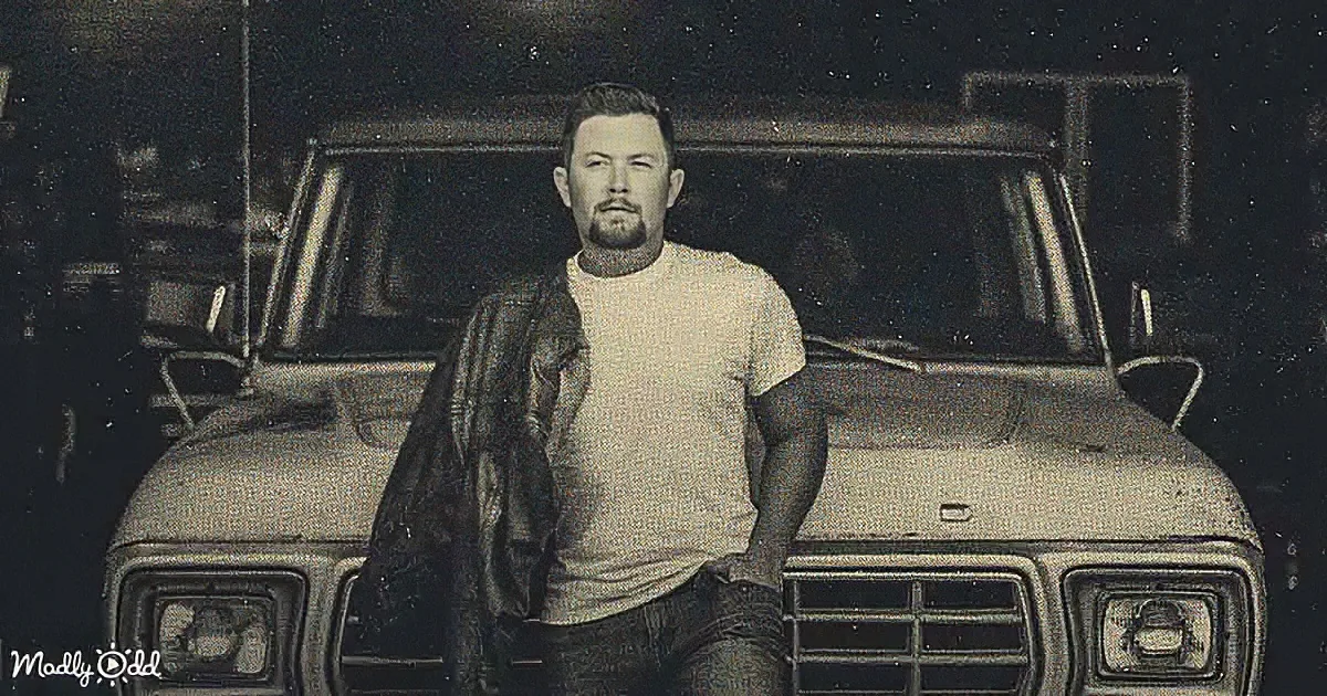A man stands in front of a vintage vehicle, wearing a white shirt and jeans, with a leather jacket draped over his shoulder.