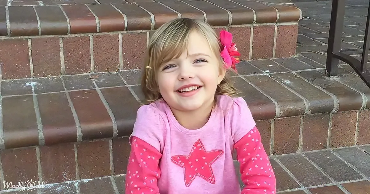 A smiling child with a pink flower in her hair, wearing a pink star-patterned shirt, sits against a brick wall.