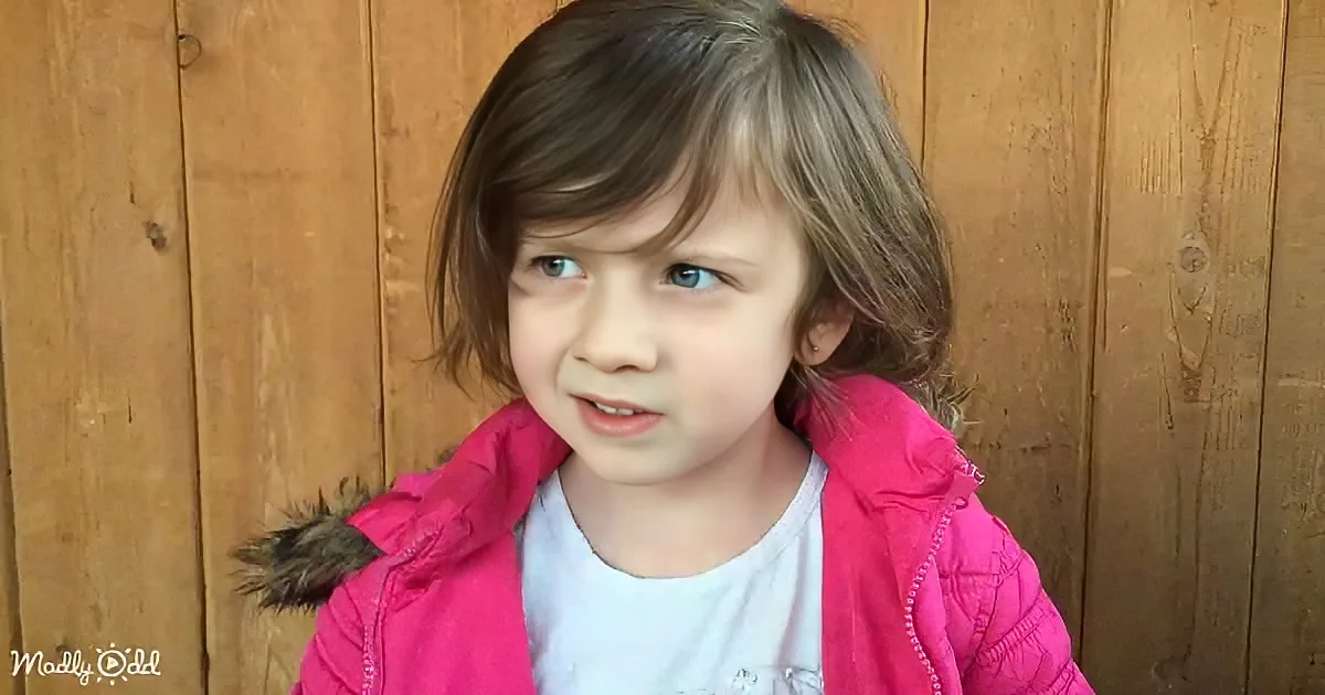 A young child with brown hair and blue eyes, wearing a pink jacket, stands against a wooden wall.