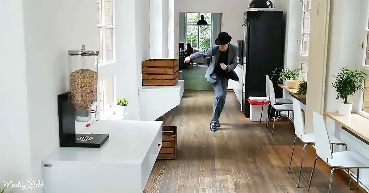 A man in a suit and hat dances in a bright, modern office space with wooden floors and white furniture.