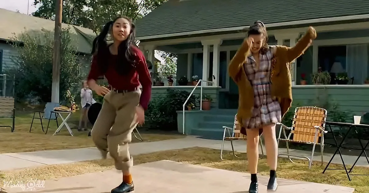 Two girls dance in a suburban backyard, one in a red sweater and khaki pants, the other in a plaid dress and brown jacket.