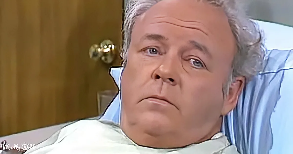 A man with gray hair lies in a hospital bed, wearing a light blue gown, with a concerned expression.