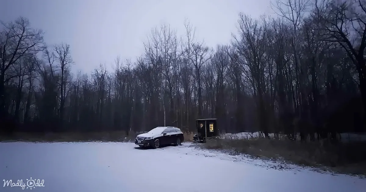 A car is parked near a small illuminated structure in a snowy, forested area.