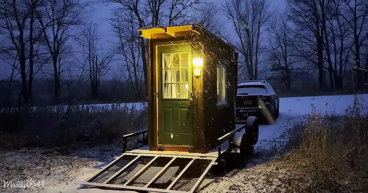 A small, illuminated house on wheels sits on a snowy path surrounded by bare trees.