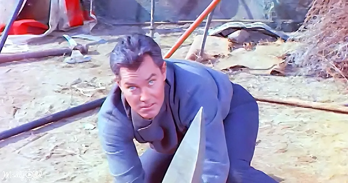 A man in a blue suit crouches on the ground, holding a knife, with a rugged outdoor setting behind him.