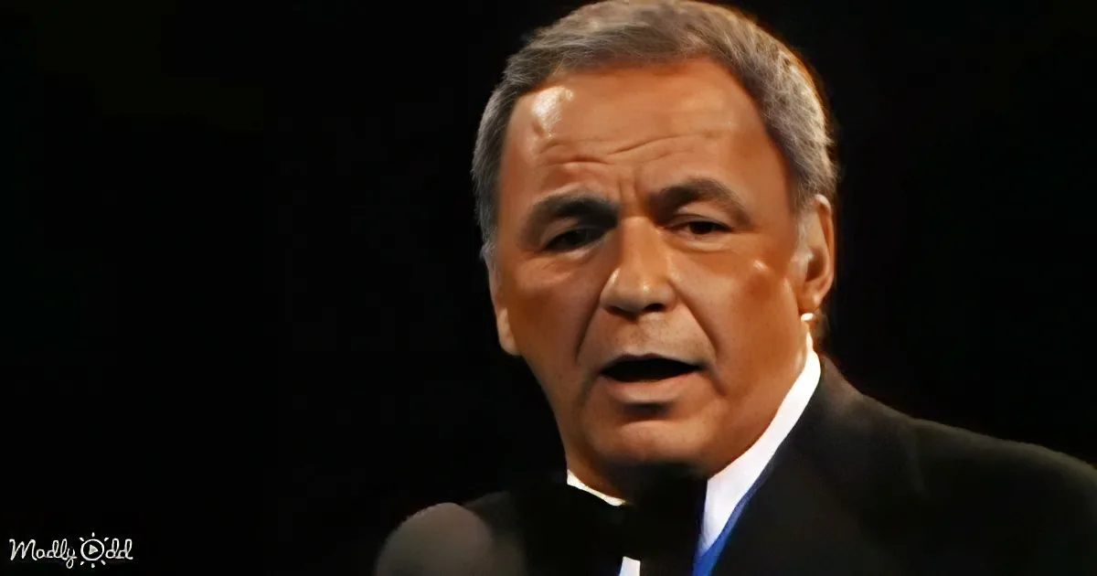 A man in a suit and bow tie sings into a microphone against a dark background.