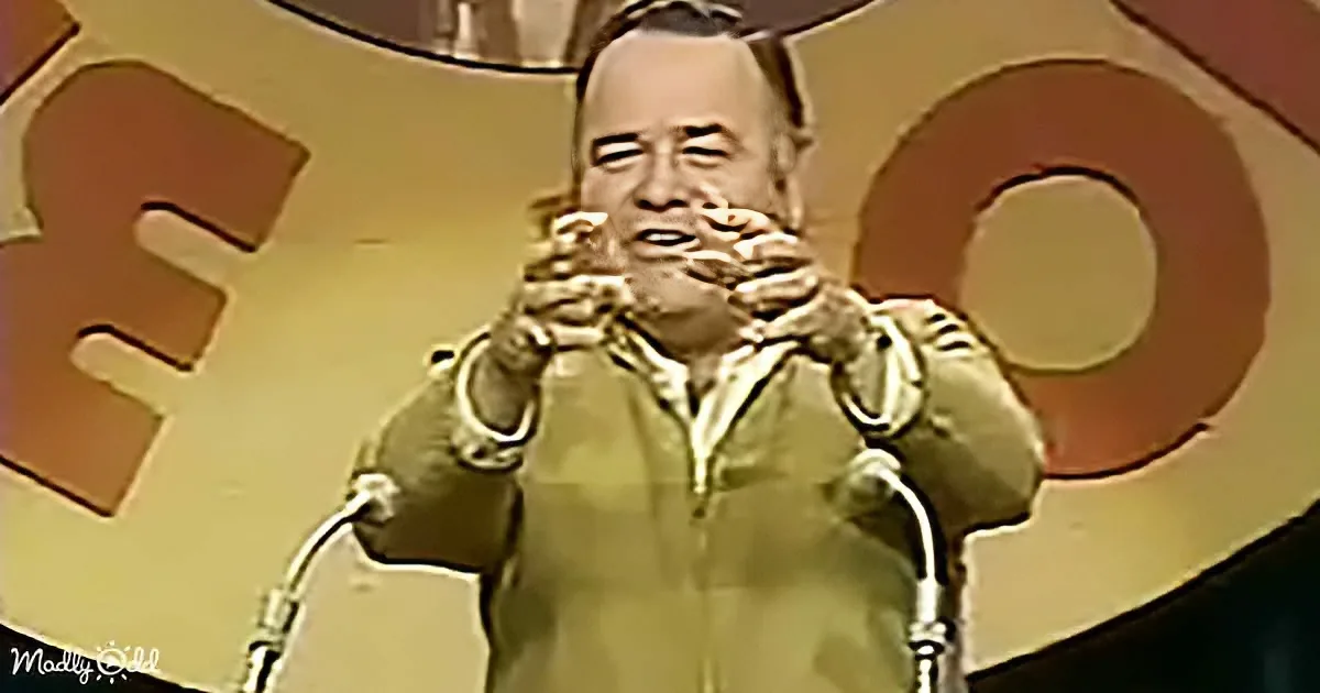 A man in a green jacket stands behind microphones, hands clasped, in front of a large "S" and "O" backdrop.