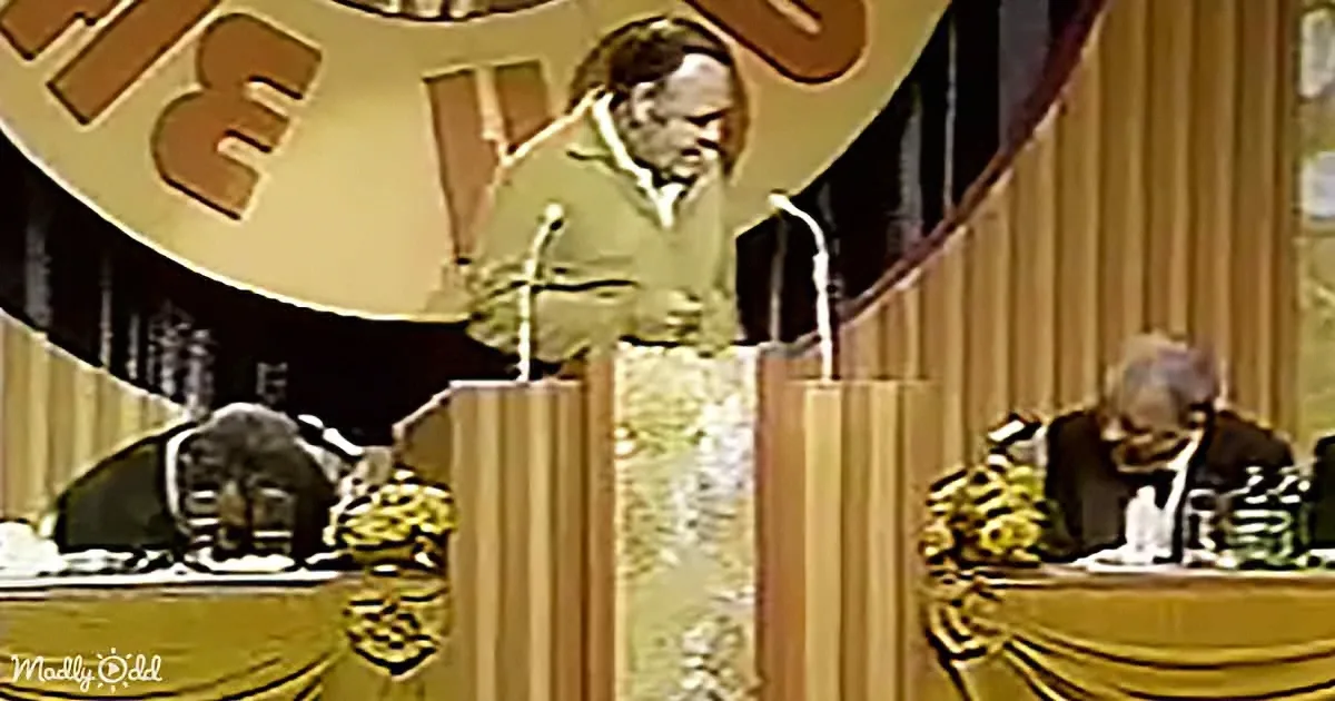 A man stands at a podium with microphones, speaking in front of a large screen displaying his image.
