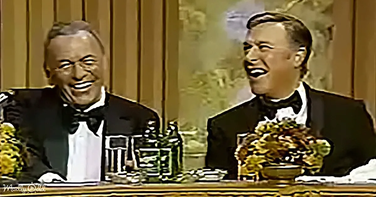 Two men in tuxedos sit at a table, laughing, with flowers and glasses in front of them.