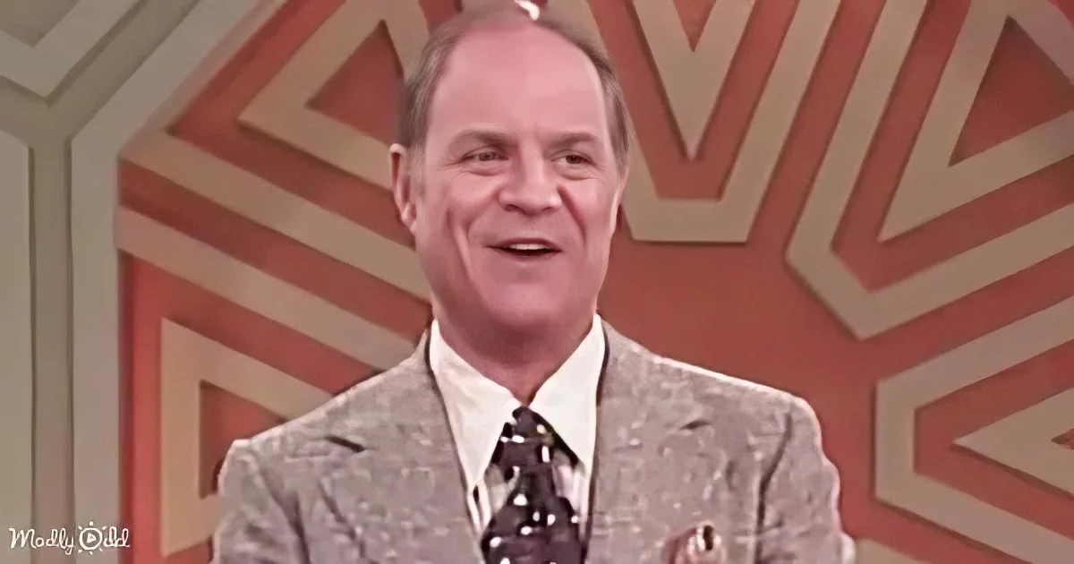 A man in a patterned suit and tie smiles on a stage with a geometric background.