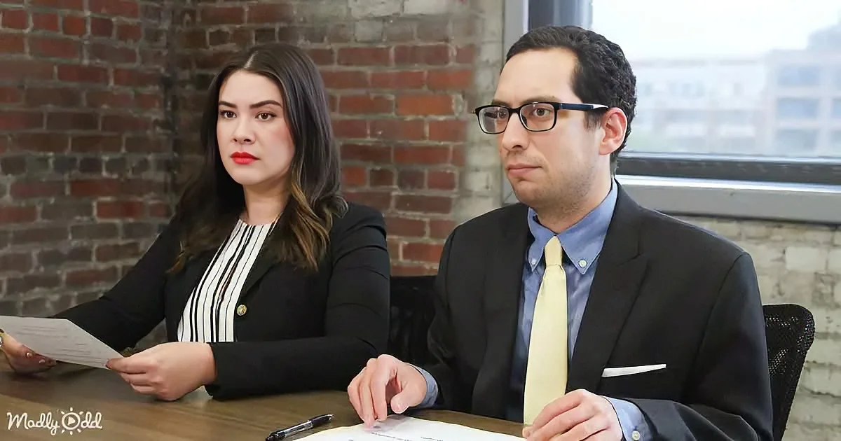 A man in a suit and tie sits at a table with a woman in a black blazer, both appearing serious.