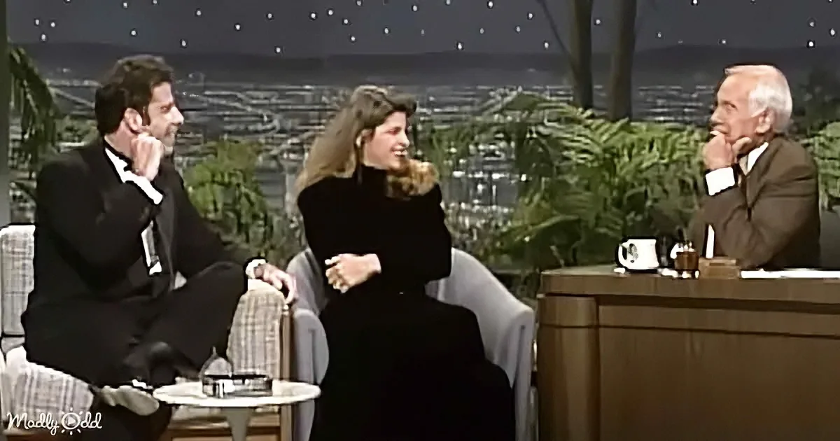 A man in a tuxedo sits on a couch, engaged in conversation with a woman and another man behind a desk.