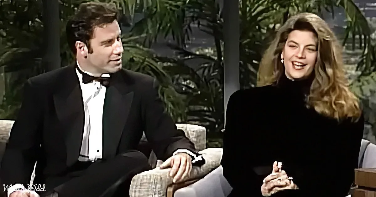A man in a tuxedo and a woman in a black top are seated on a couch, smiling and laughing.