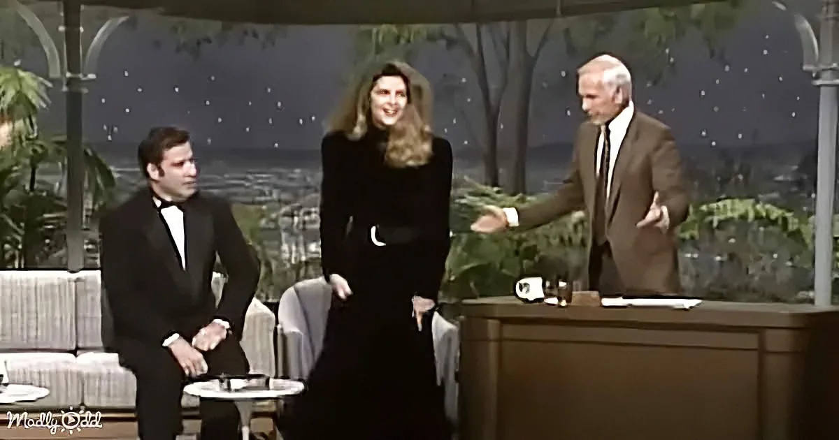 A woman in a black dress stands between two men in suits on a talk show set.