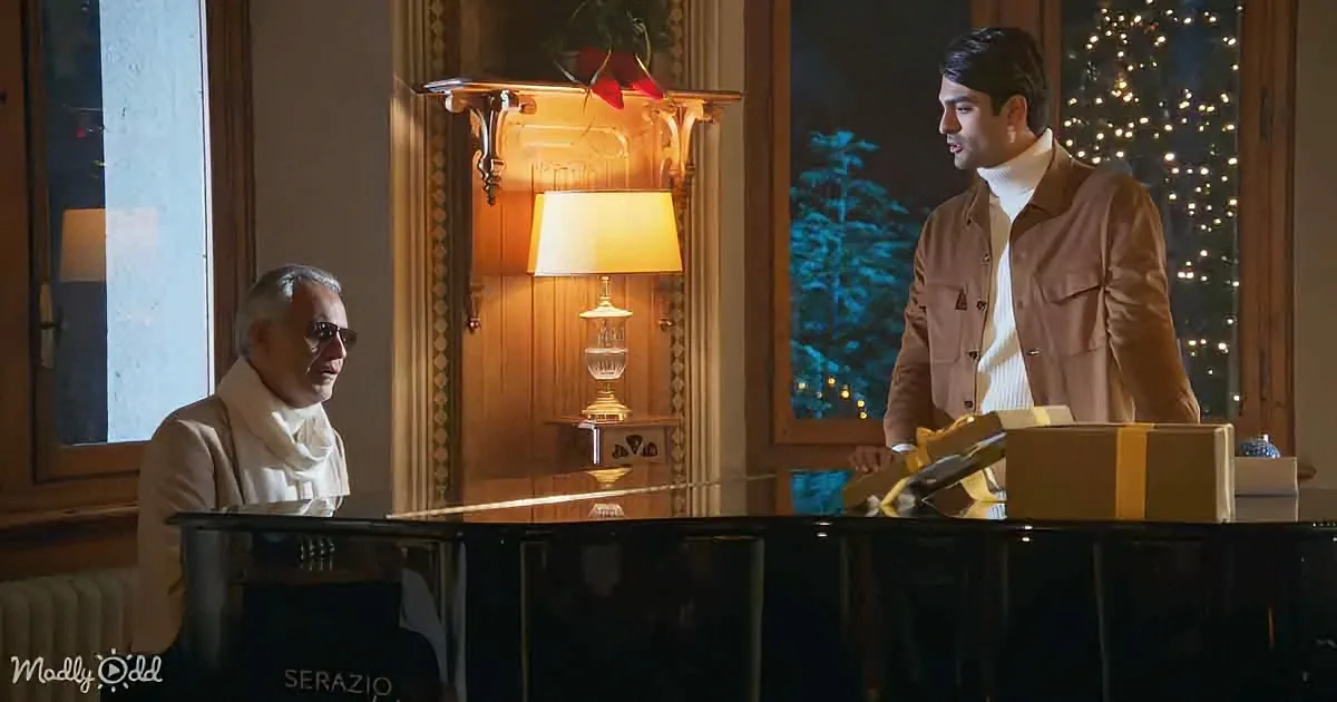 A man in a brown jacket stands beside a piano, holding a gift, while another man in a white scarf sits at the piano.
