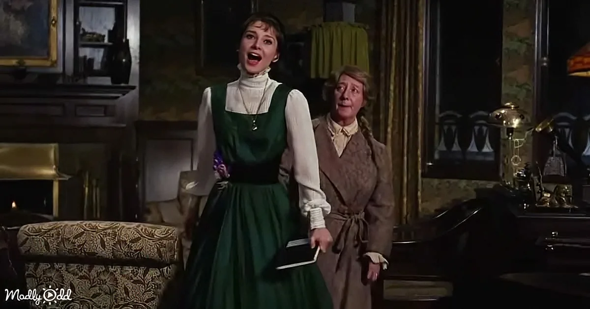 Audrey Hepburn in a green dress stands with a surprised expression, holding a book, in a dimly lit room with another...