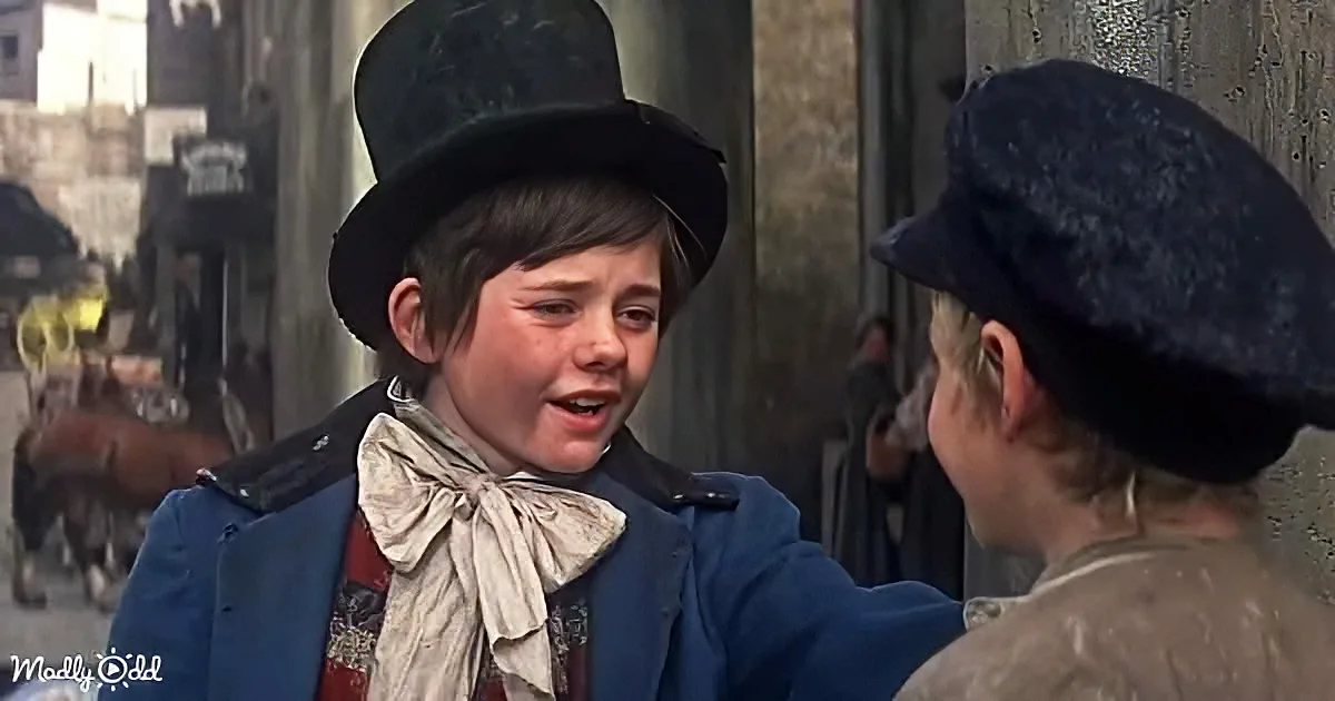 A young boy in a top hat and blue coat is speaking to another boy in a dark cap, set against a backdrop of a bustling...