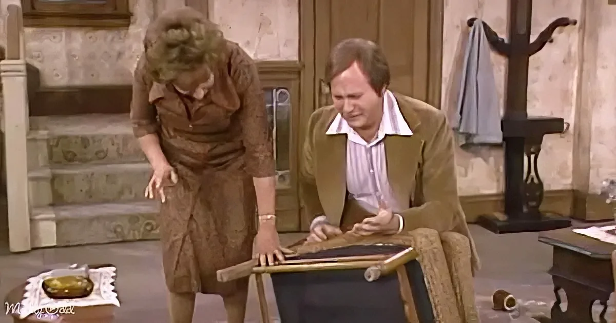 A woman in a brown dress bends over, while a man in a brown jacket leans on a table, both in a room with stairs and a...