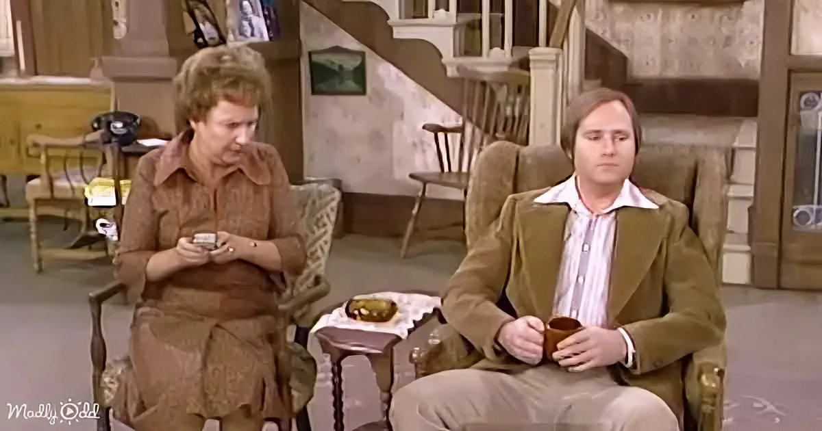 A woman in brown sits in a chair, holding a small object, while a man in a brown suit sits across from her, holding a mug.