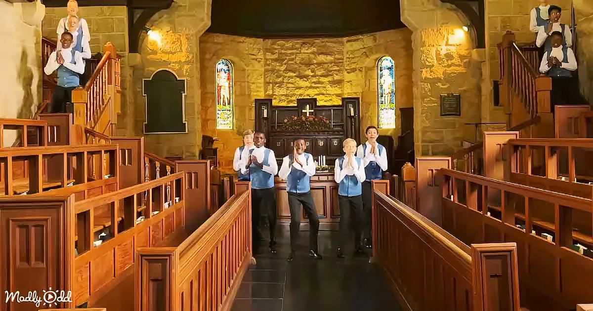 A group of five people in blue vests and white shirts sing in a church with wooden pews and stained glass windows.
