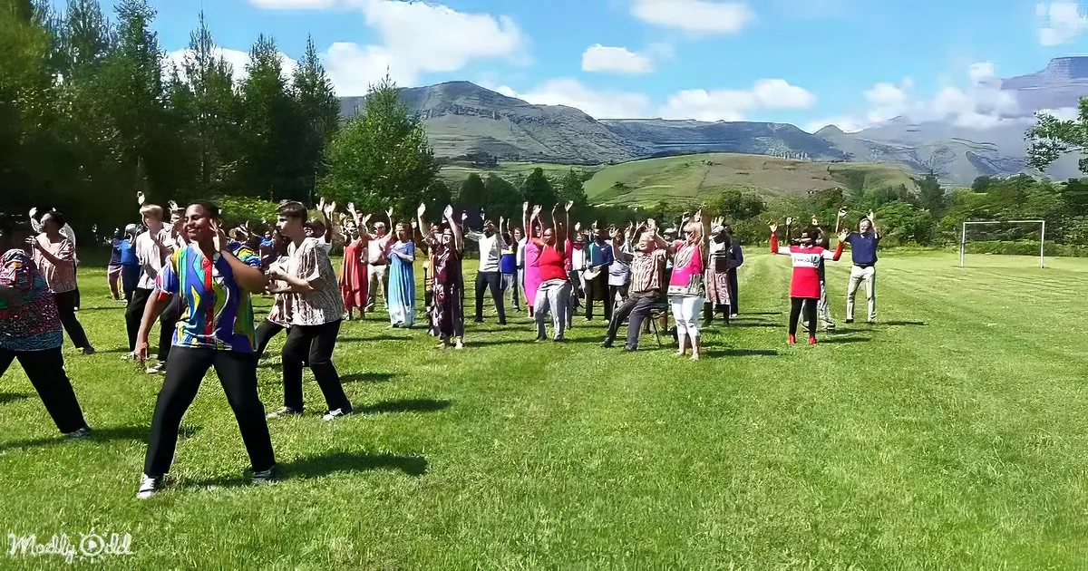 A group of people in colorful attire is dancing joyfully on a grassy field with mountains in the background.