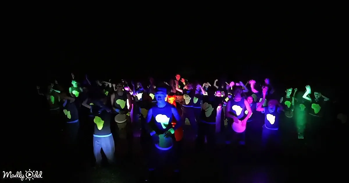 A group of people in glowing outfits dances under blacklight, creating vibrant colors.